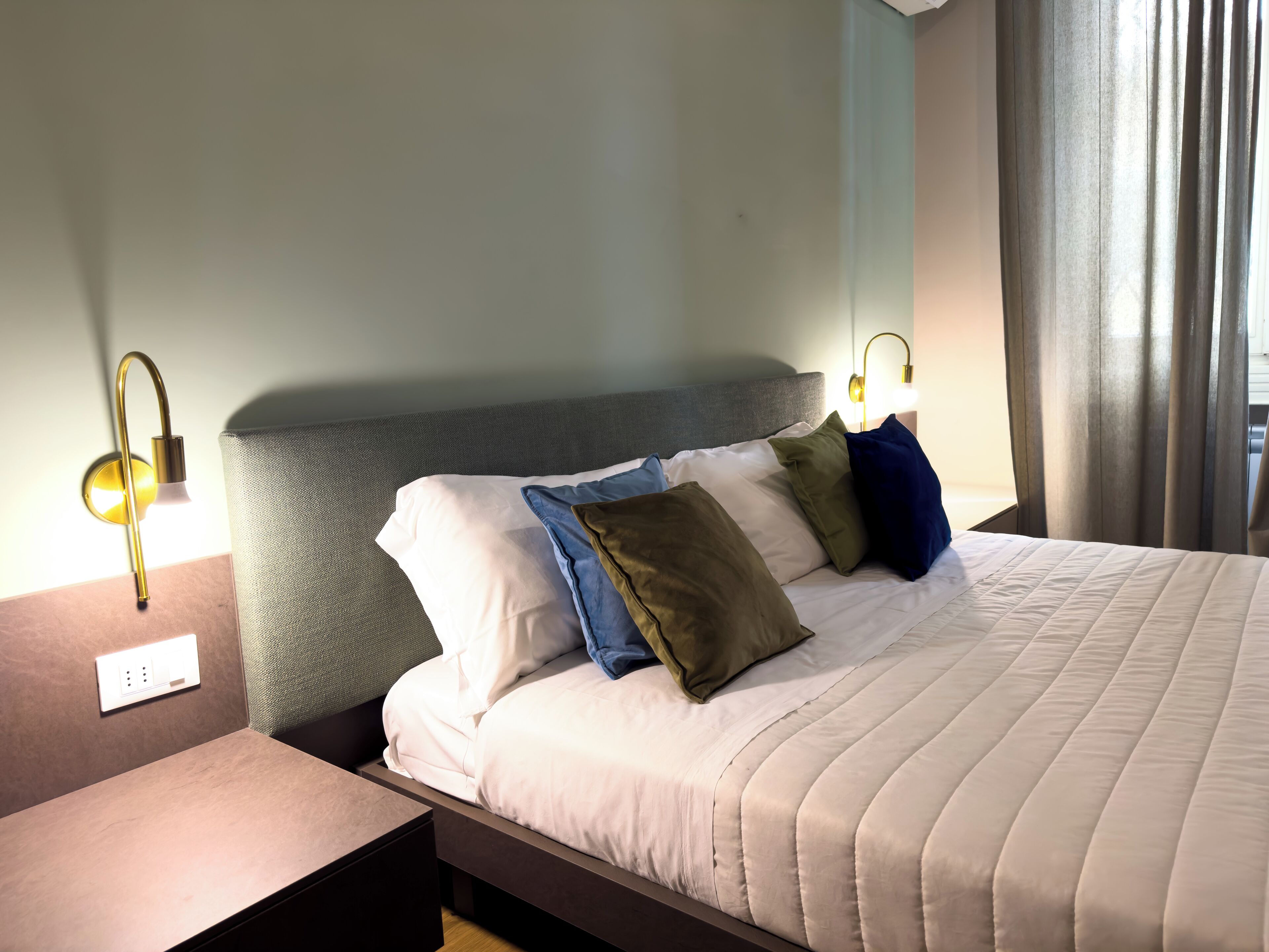 Cama bem arrumada com edredom branco e várias almofadas, cabeceira estofada cinza e mesa de cabeceira com abajur.
