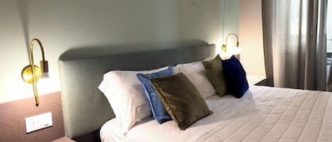 Cama bem arrumada com edredom branco e várias almofadas, cabeceira estofada cinza e mesa de cabeceira com abajur.