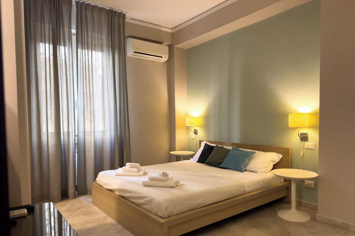 A modern hotel room with a wooden bed, two bedside tables, an air conditioning unit, and a large window with curtains.