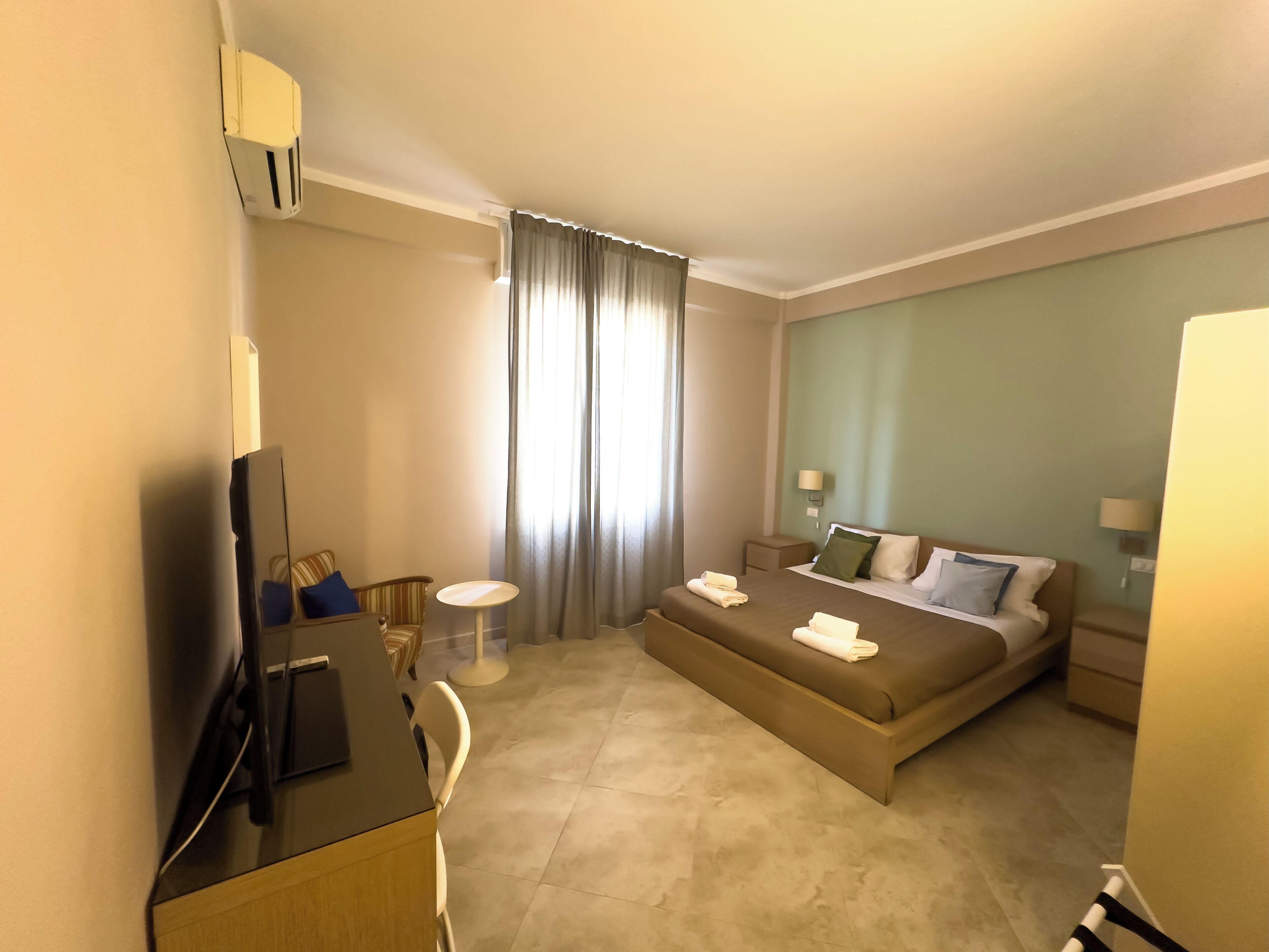 A hotel room with a bed, a desk, a chair, a TV, a lamp, and a window with curtains.