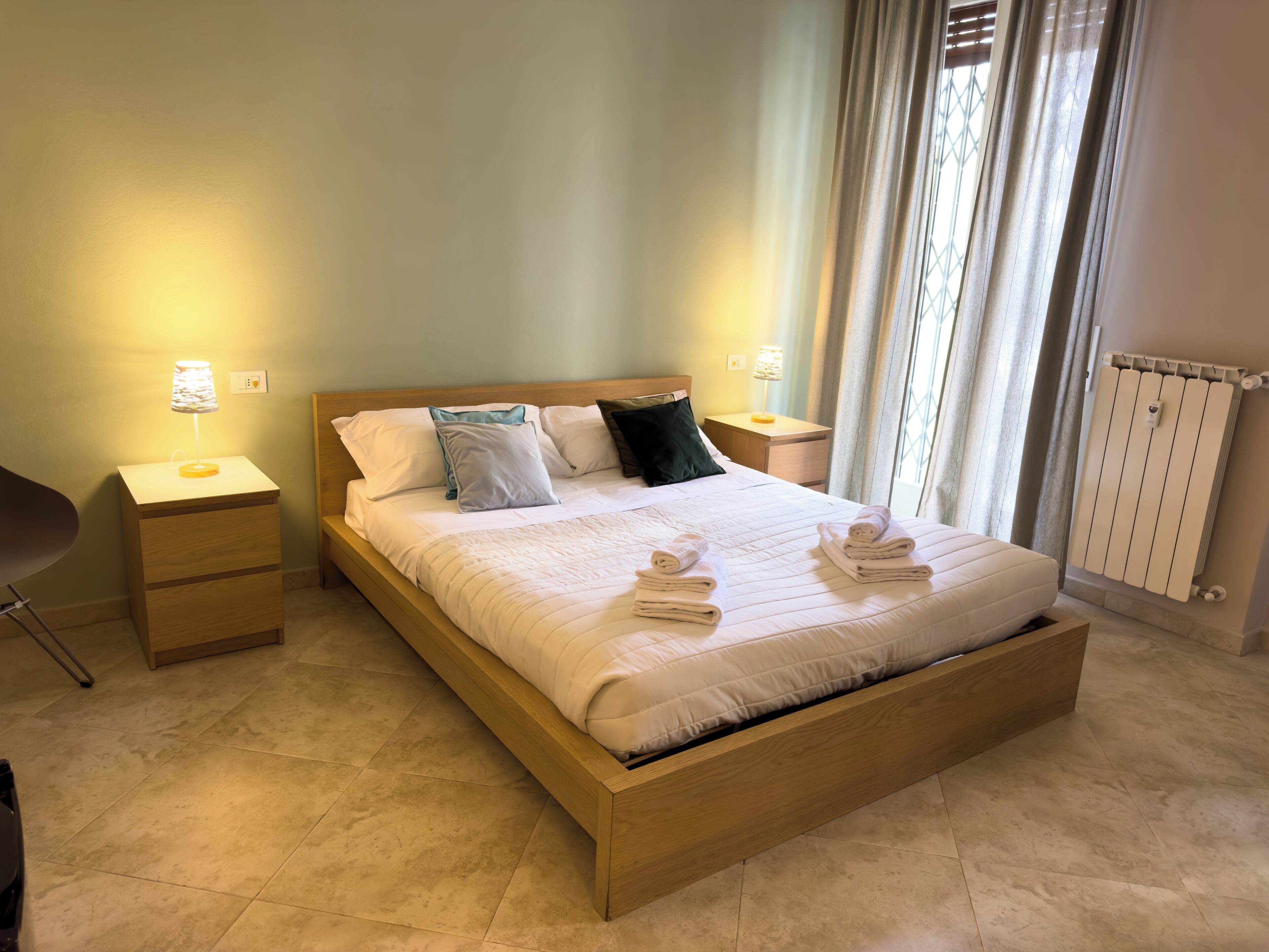 A neatly made bed with a wooden frame and a quilted white duvet. There are two bedside tables with lamps, a chair, and a radiator.