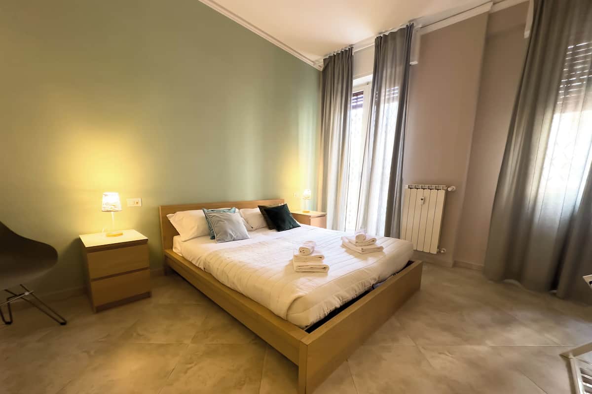 A neatly made bed with a wooden headboard, a bedside table with a lamp, and a radiator on the wall.