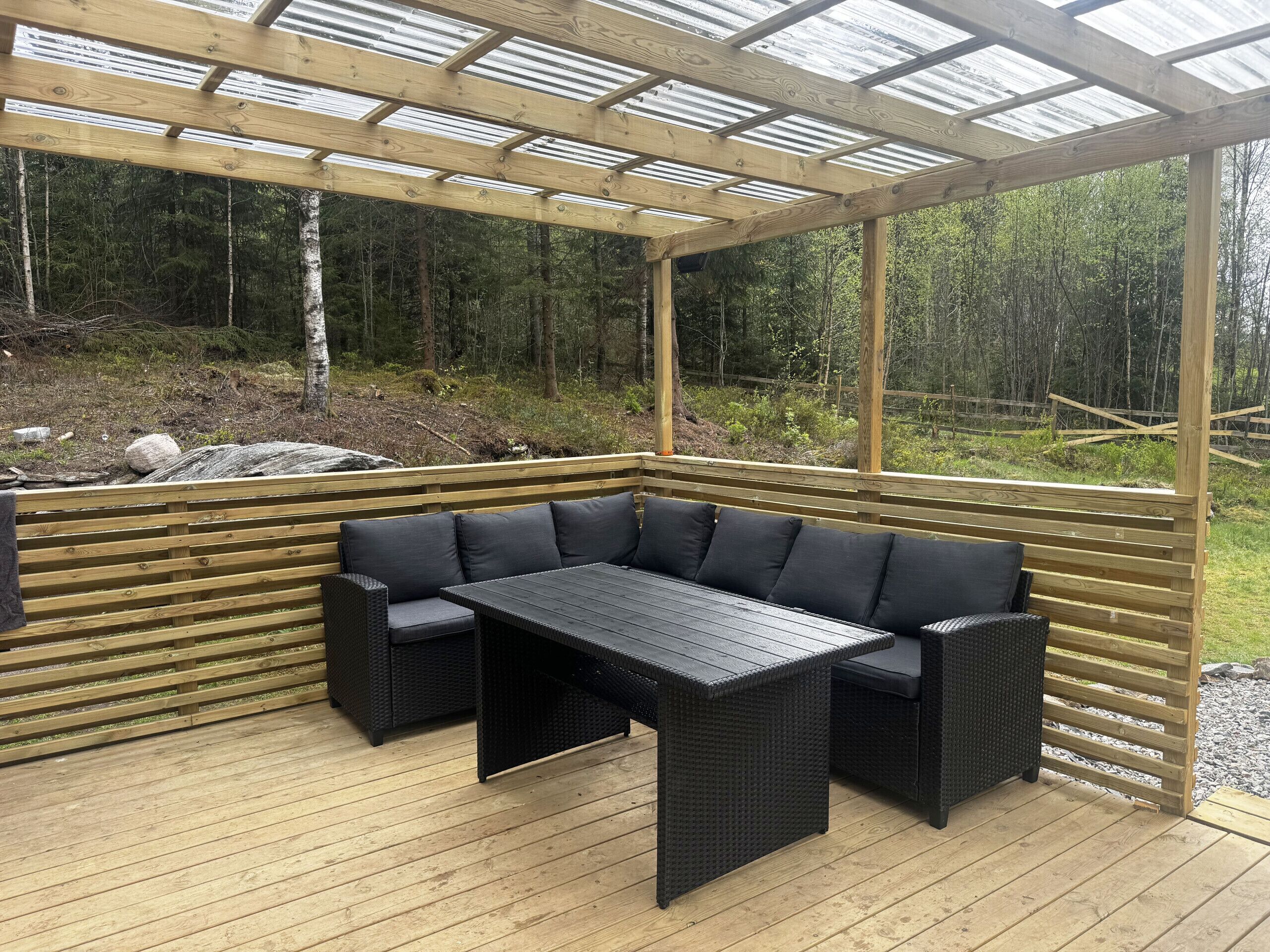 Un espacio al aire libre cubierto con mesa y sillas de madera, rodeado por una terraza de madera y paneles de vidrio.
