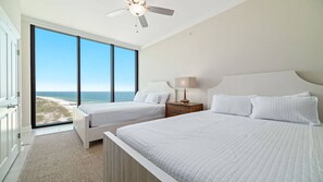 Habitación de hotel con dos camas, un ventilador de techo, una mesita de noche con lámpara y vista a la playa.
