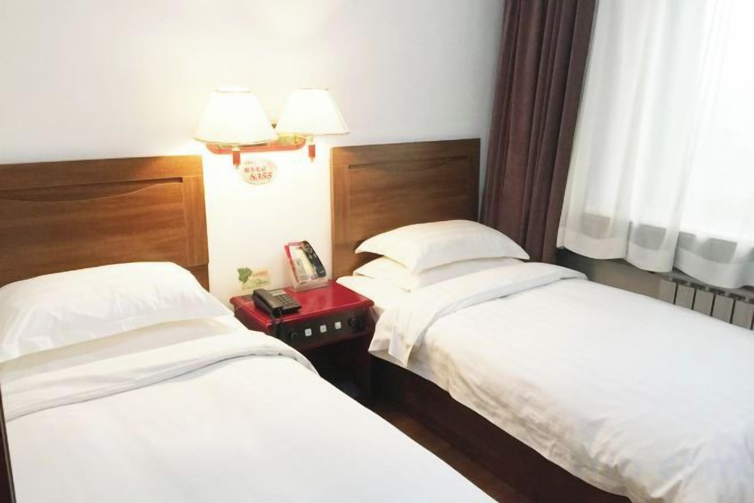 Habitación de hotel con dos camas individuales, cabecero de madera, mesita de noche con lámpara y ventana con cortinas.