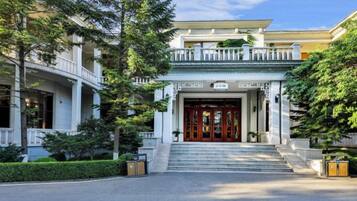 Entrada de un hotel con puertas dobles, escalones de acceso y un pequeño jardín con un banco.