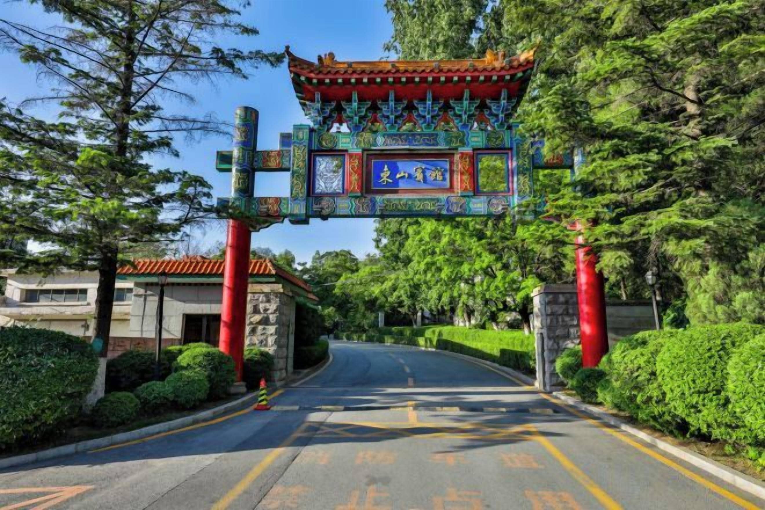 A traditional Chinese gate with intricate designs, leading to a road flanked by neatly trimmed bushes and trees.