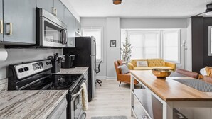 Een moderne keuken met een fornuis, magnetron en koelkast. Er is een zithoek met een houten tafel en een gele bank.