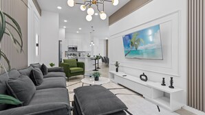 A modern living room with a grey sofa, a green armchair, a white entertainment unit, and a large TV on the wall. - Prime Location: Townhome with Pool & Game Room! (Orlando)