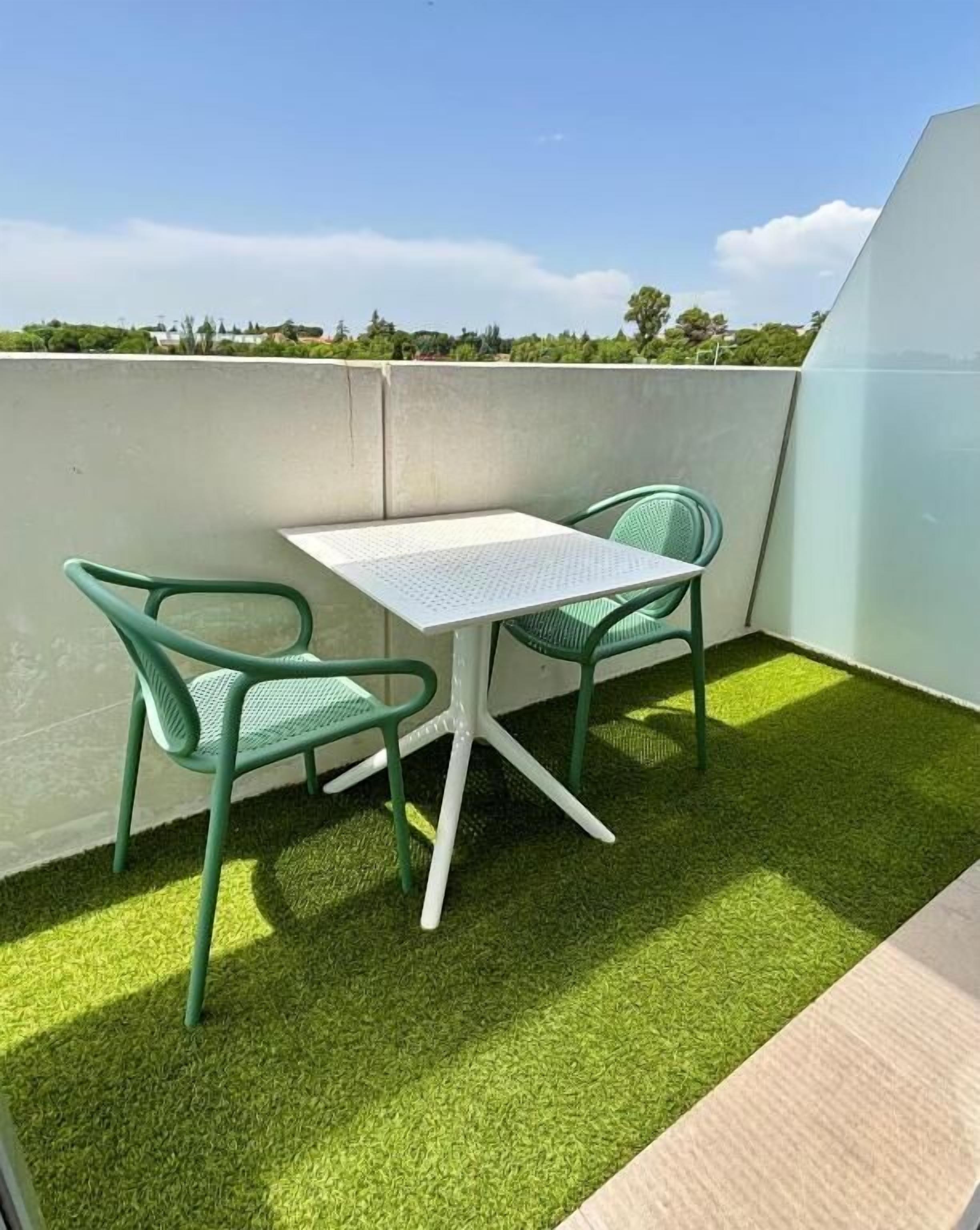 A small outdoor balcony with a table and two chairs, artificial grass flooring, and a view of trees and buildings in the background.