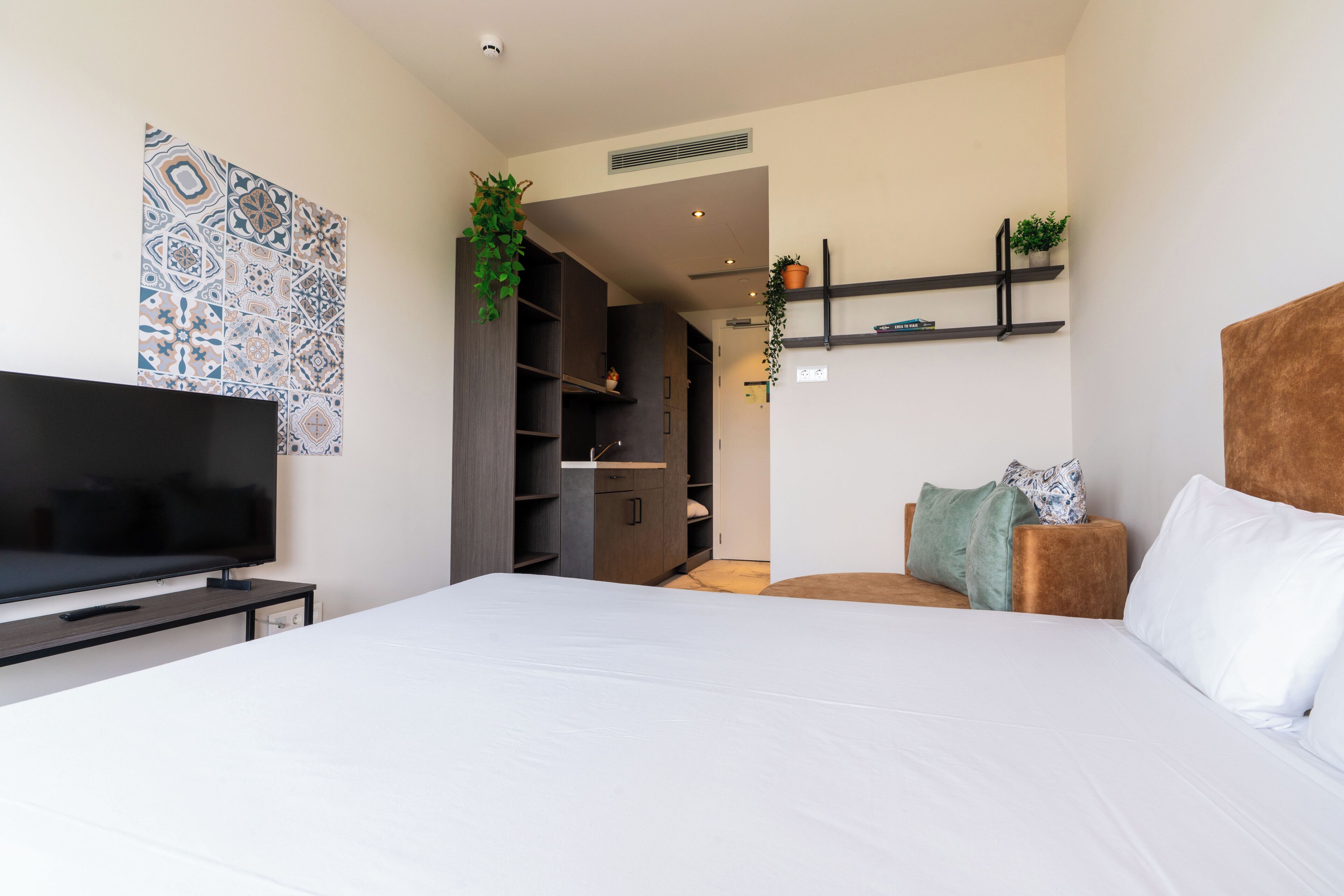 A modern bedroom with a large bed, a television, a wardrobe, and a small seating area with cushions.