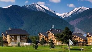 A row of wooden cabins with stone foundations, set against a backdrop of snow-capped mountains and a clear sky.