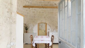 A rustic stone-walled room with a wooden table and chairs, a white cabinet, and a large window with a view. - Trullo Puspo by Wonderful Italy (Carovigno)