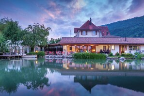 A lakeside hotel with a reflection in the water, a covered outdoor seating area, and a mountain backdrop. - Gasthof zum Reh - Self-Check-in (Herbetswil)