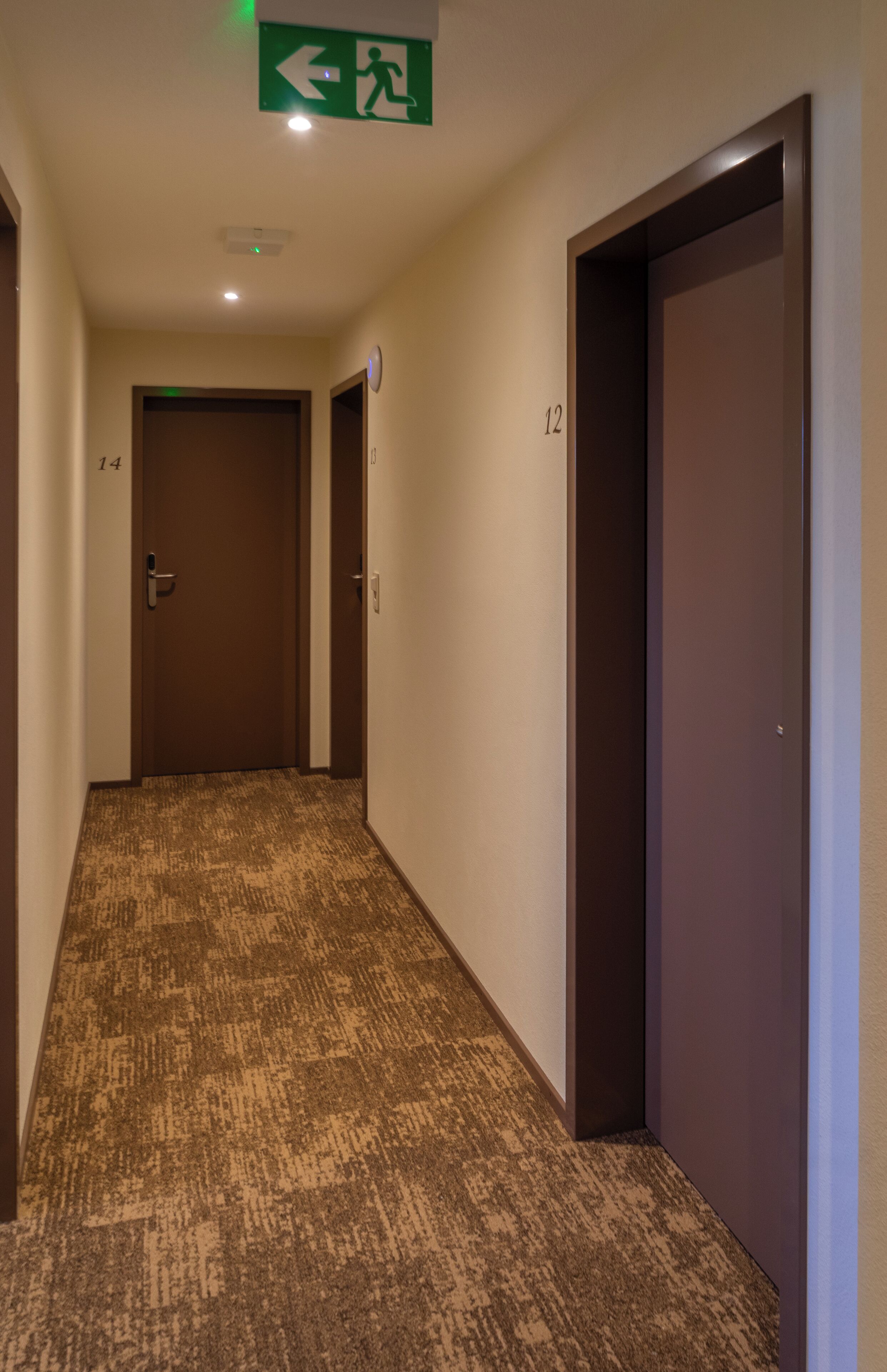 Um corredor de hotel com pisos carpetados, portas com os números 12 e 14, e um sinal de saída de emergência.