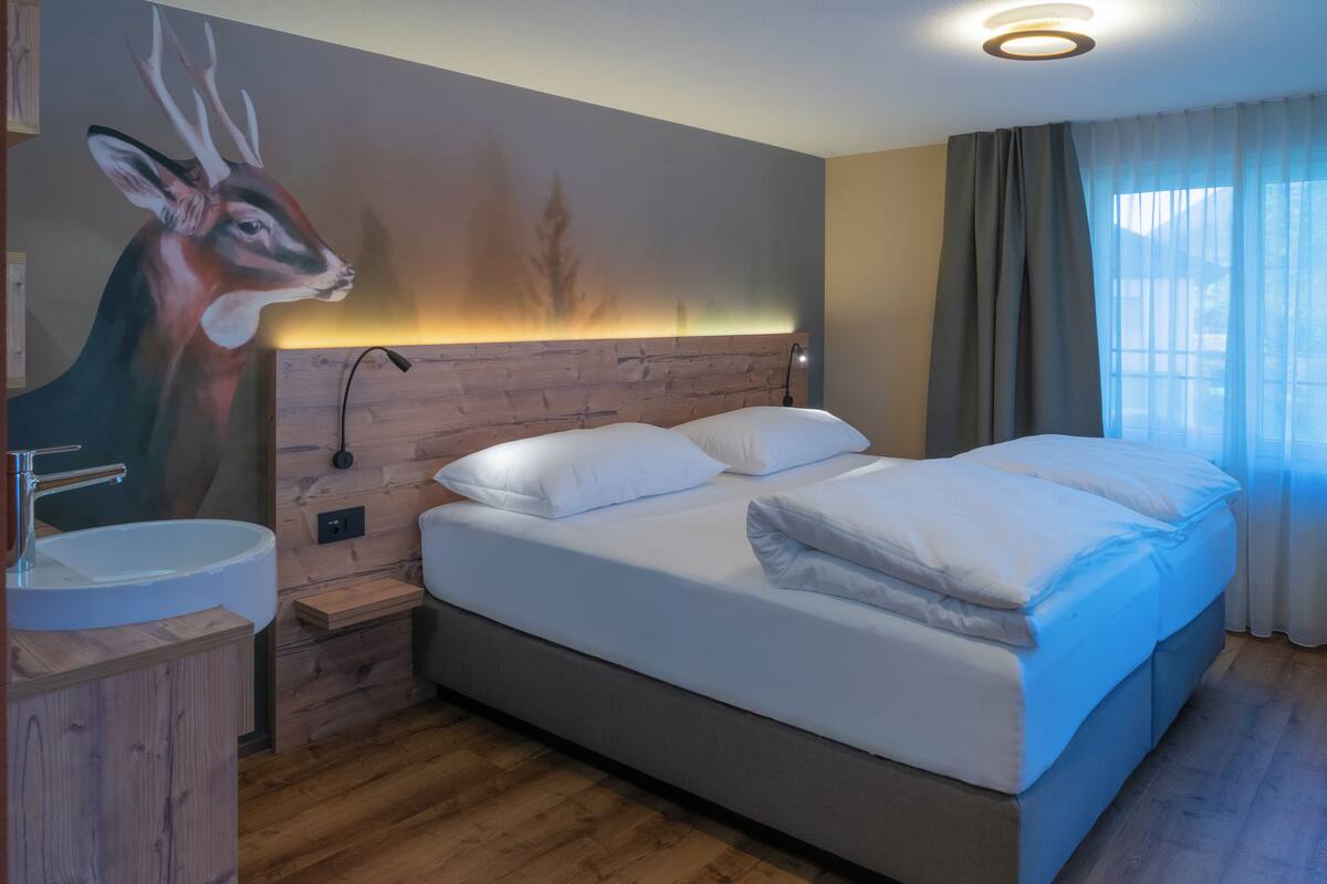 A modern hotel room with a wooden headboard, a large bed with white linens, a bedside table, and a wall-mounted painting of a deer.