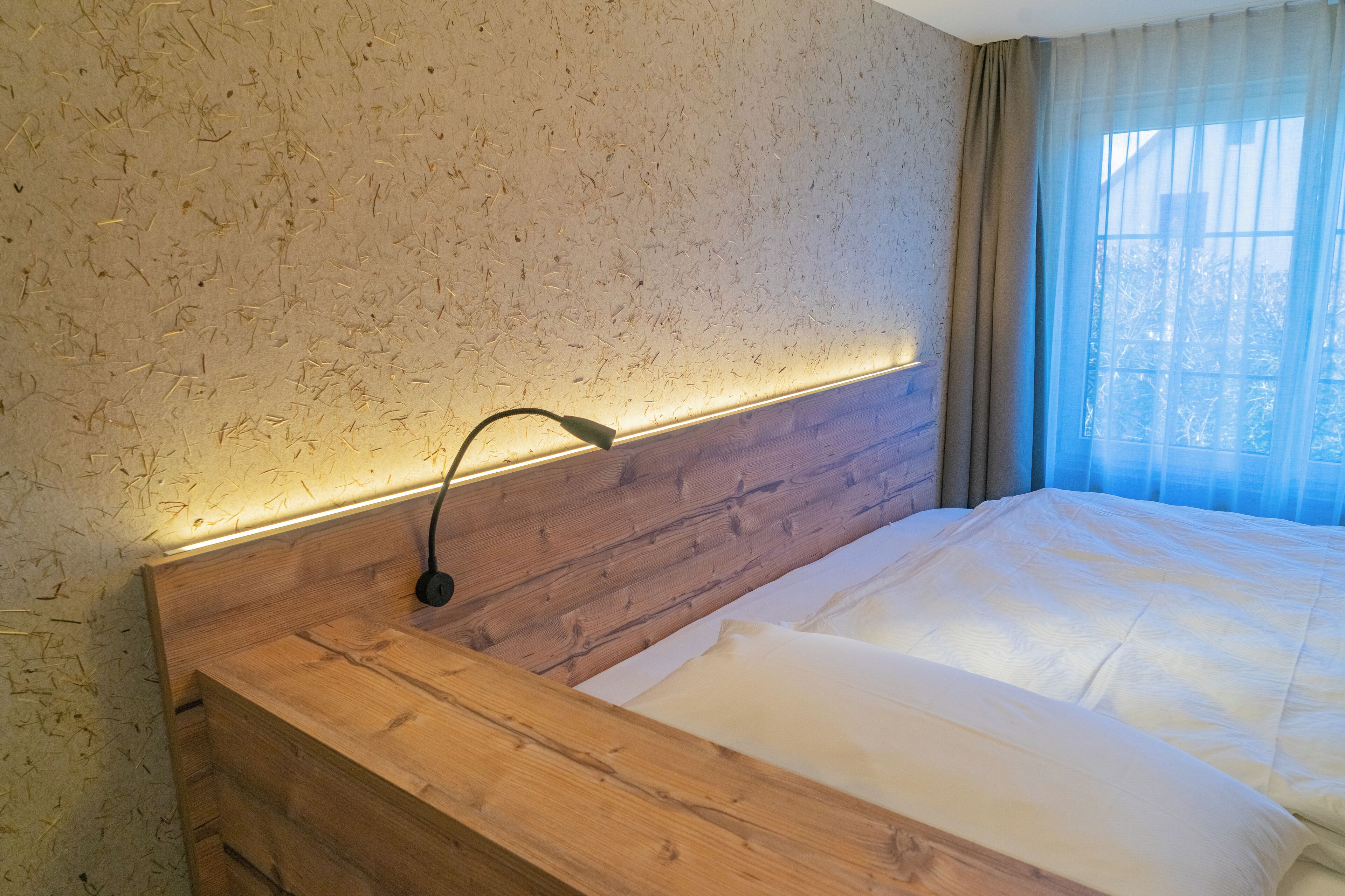 Ein hölzerner Kopfseitenverzierer mit integrierter Beleuchtung, ein ordentlich bezogenes Bett mit weißer Bettwäsche und ein Fenster mit durchsichtigen Vorhängen.