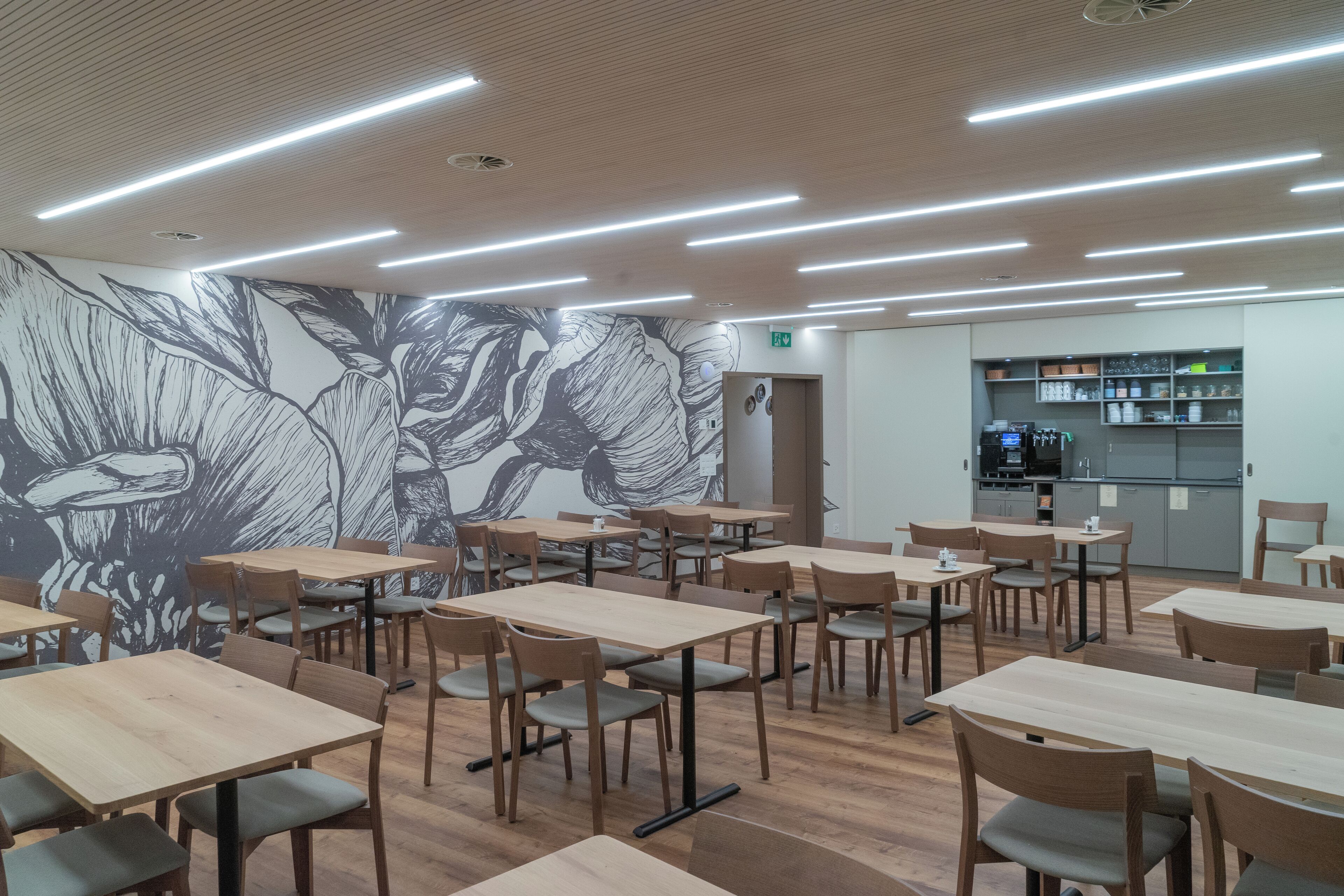 A modern cafe with wooden tables and chairs, a large floral mural on the wall, and a kitchen area in the background.