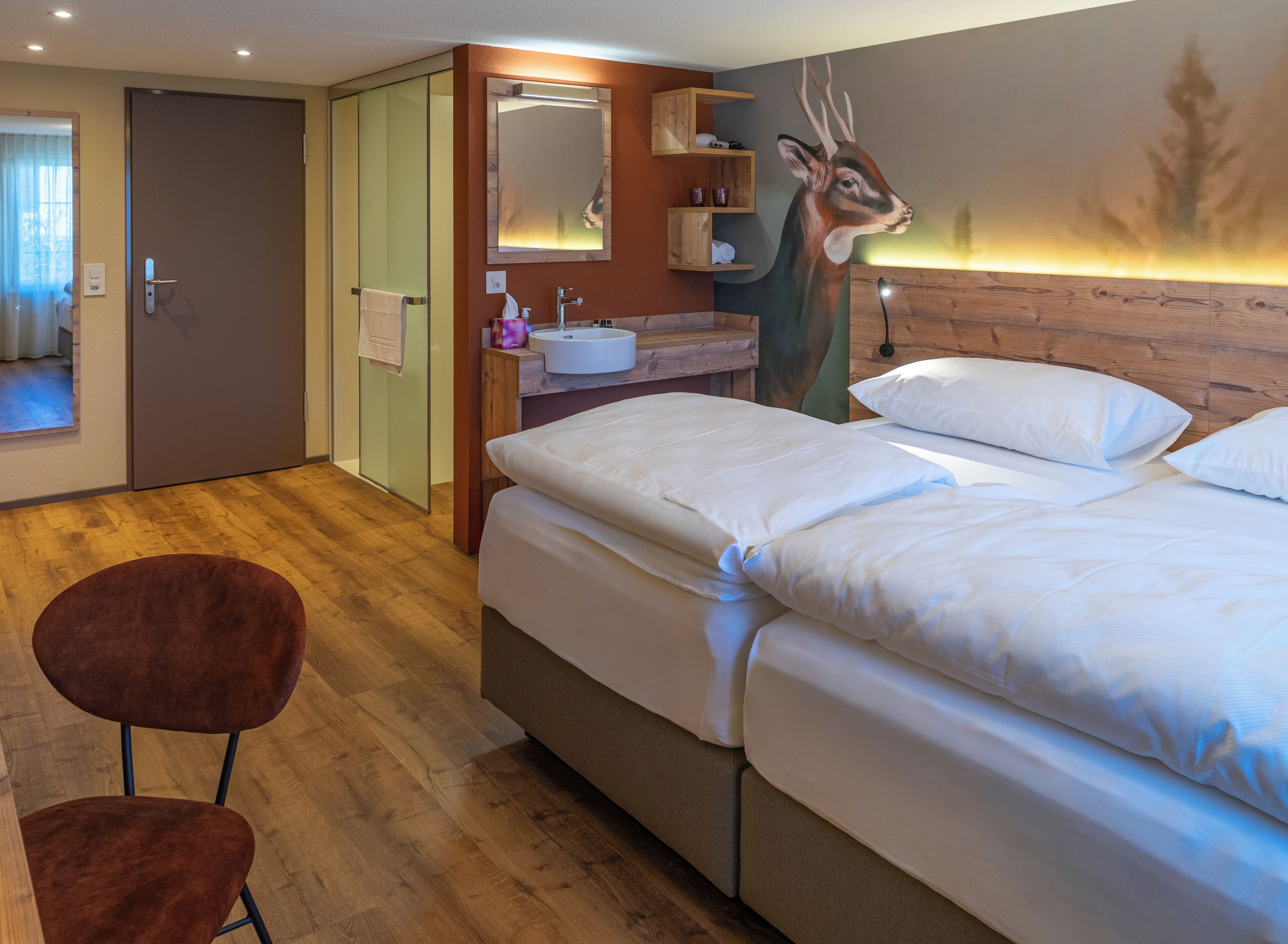 Una habitación de hotel con cama, una silla, baño con lavamanos y espejo, y un cuadro de un ciervo en la pared.