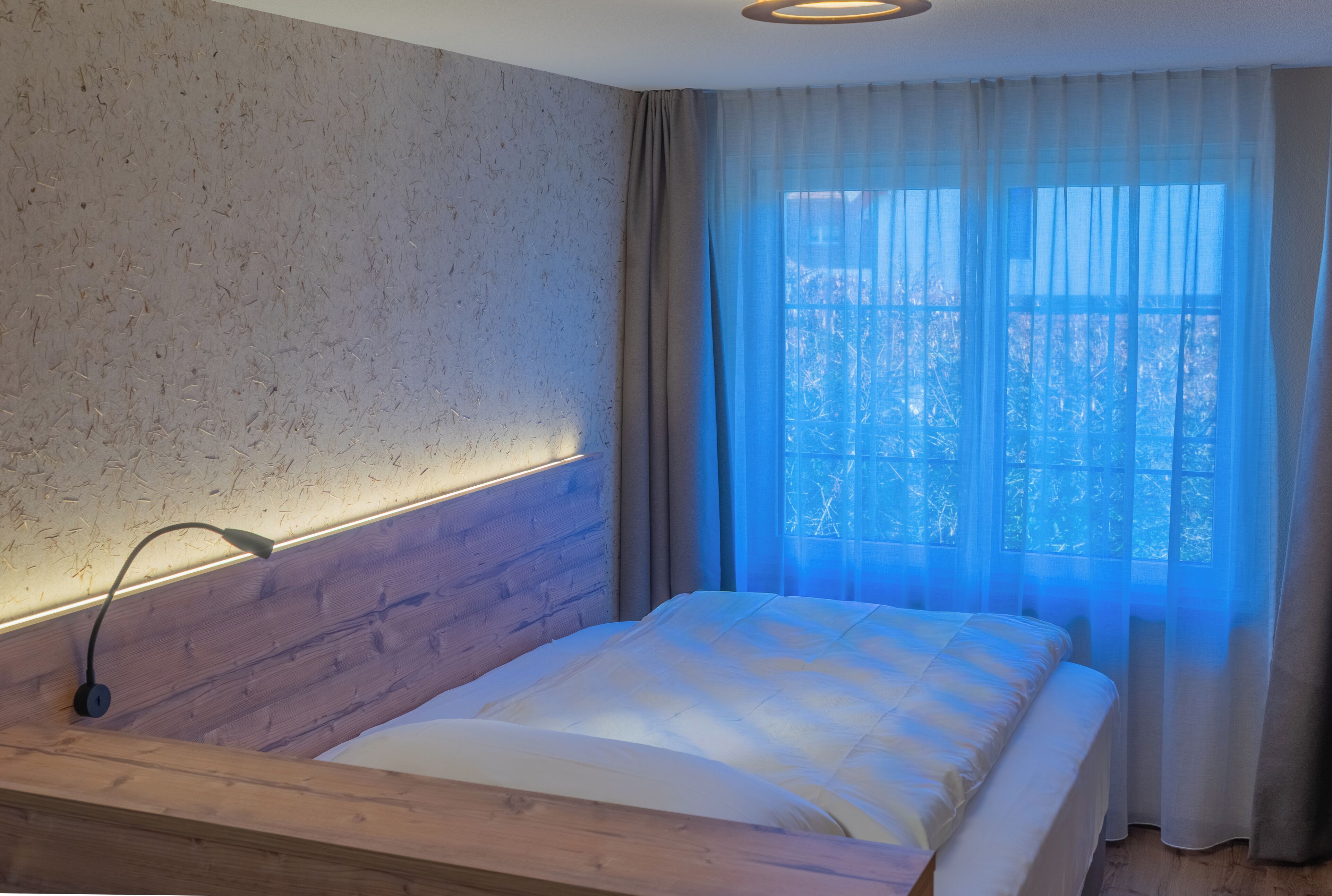A bedroom with a wooden headboard, a bed with white bedding, and a window with sheer curtains.