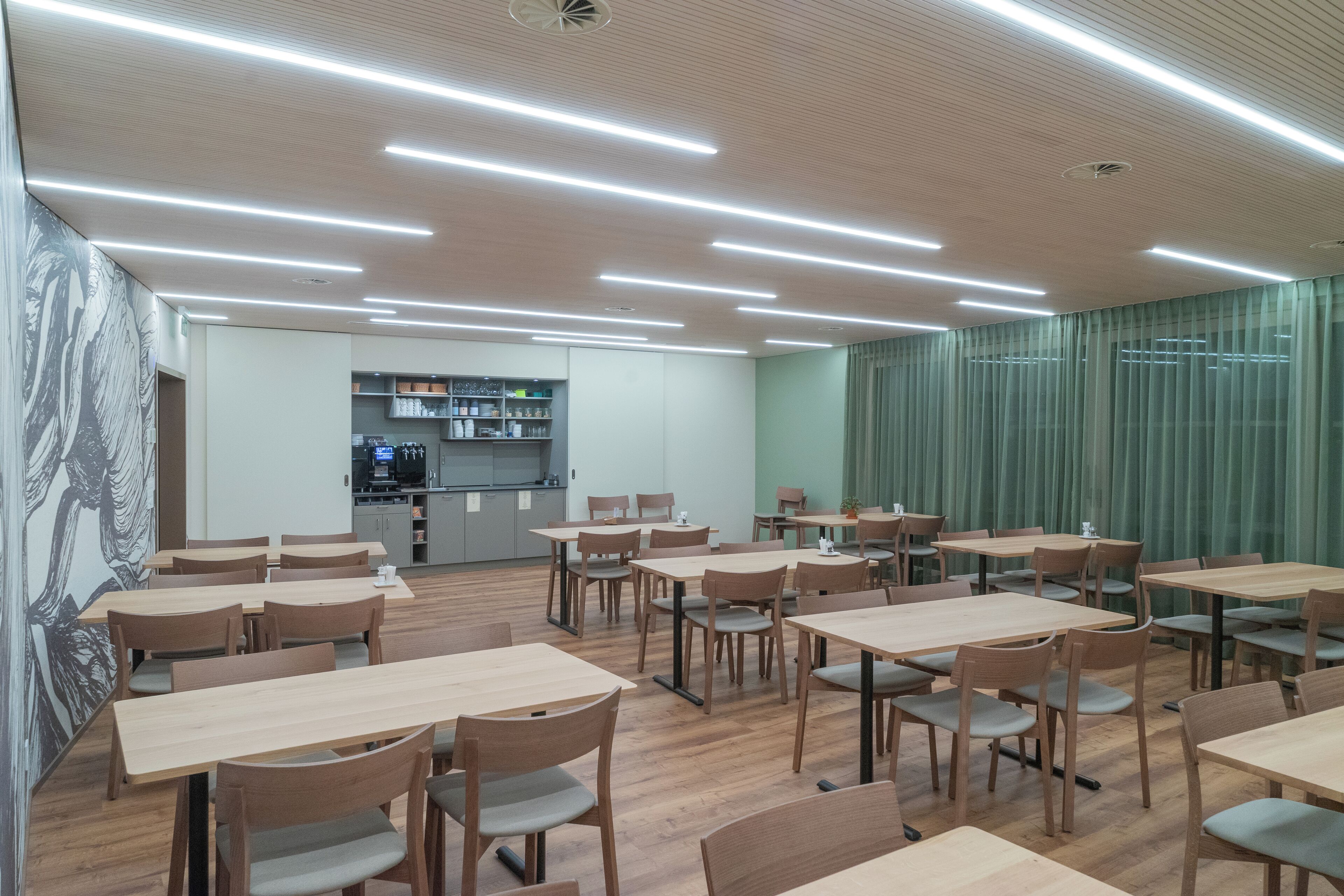 Un comedor moderno con mesas y sillas de madera, una cocina al fondo y un muro con cortinas verdes.