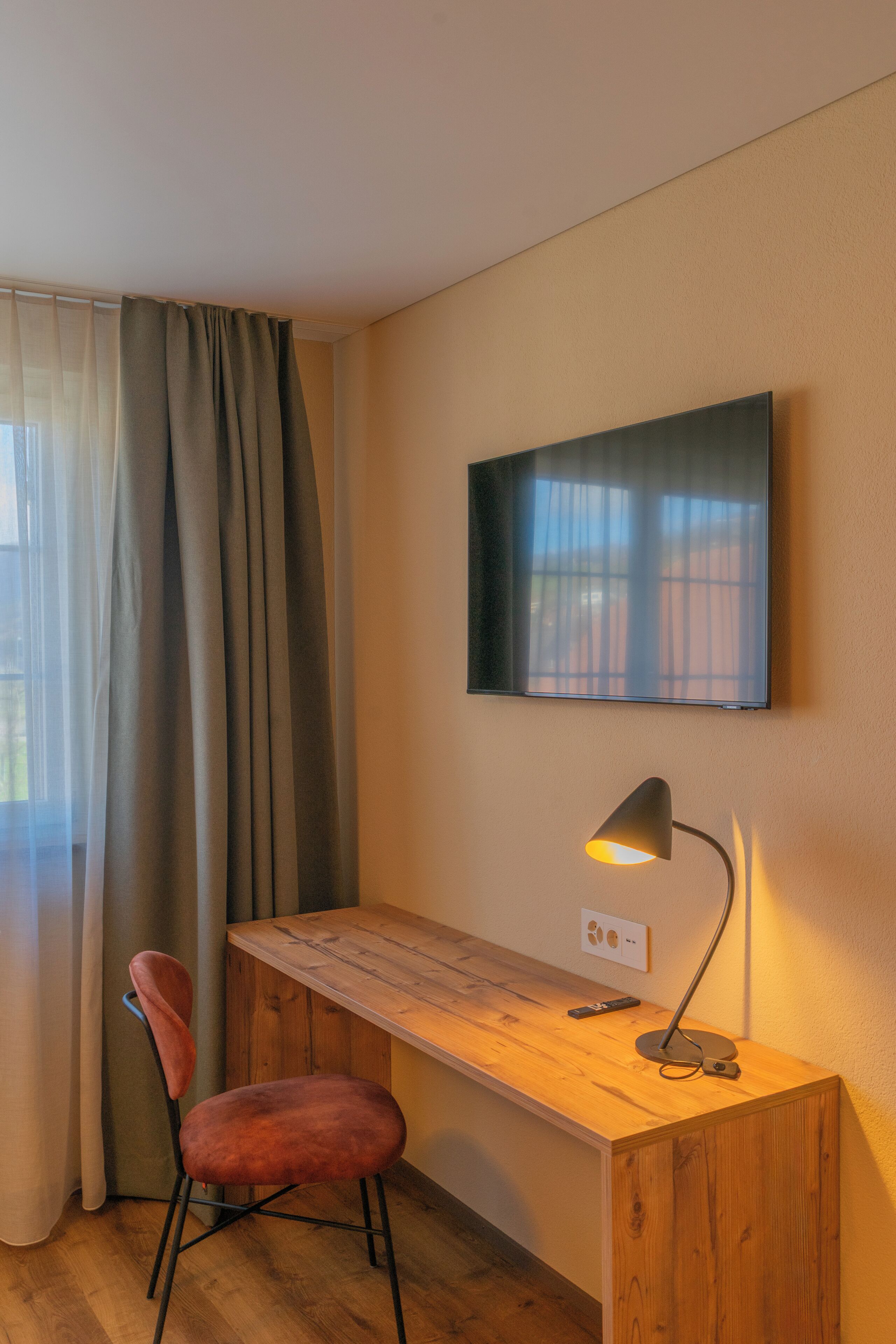 Una habitación con un escritorio de madera, una silla roja, un televisor montado en la pared y una lámpara sobre el escritorio.