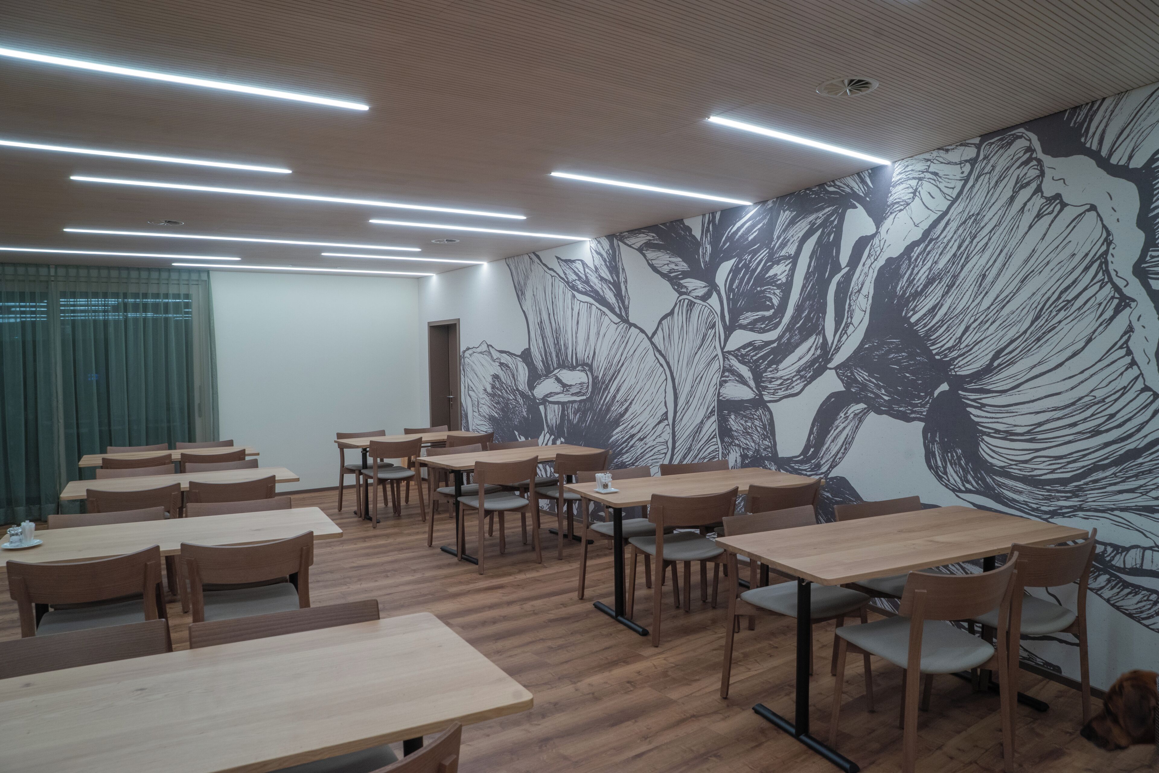 A modern dining area with wooden tables and chairs, a large floral mural on the wall, and recessed ceiling lighting.