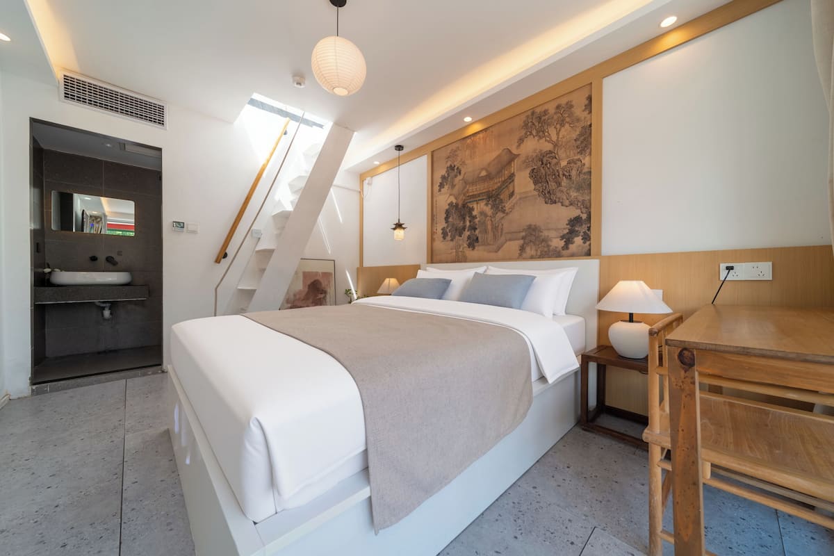 A modern hotel room with a large bed, a wooden desk, a nightstand, a wall-mounted painting, and a staircase.