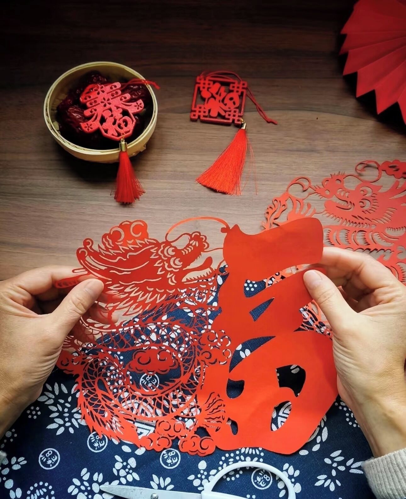 Hands holding a red paper cut-out, with a bowl of red items and a red tassel in the background.