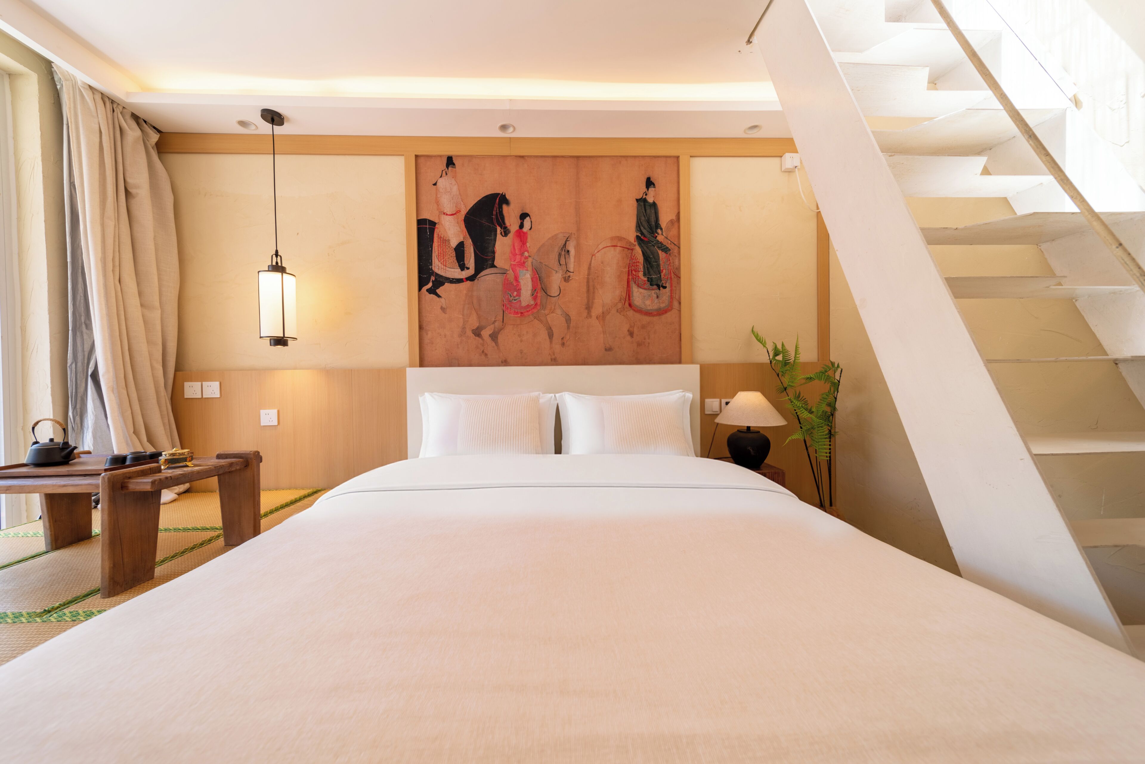 Habitación de hotel con una cama grande, un cuadro en la pared, una escalera y una mesita de noche con una lámpara.