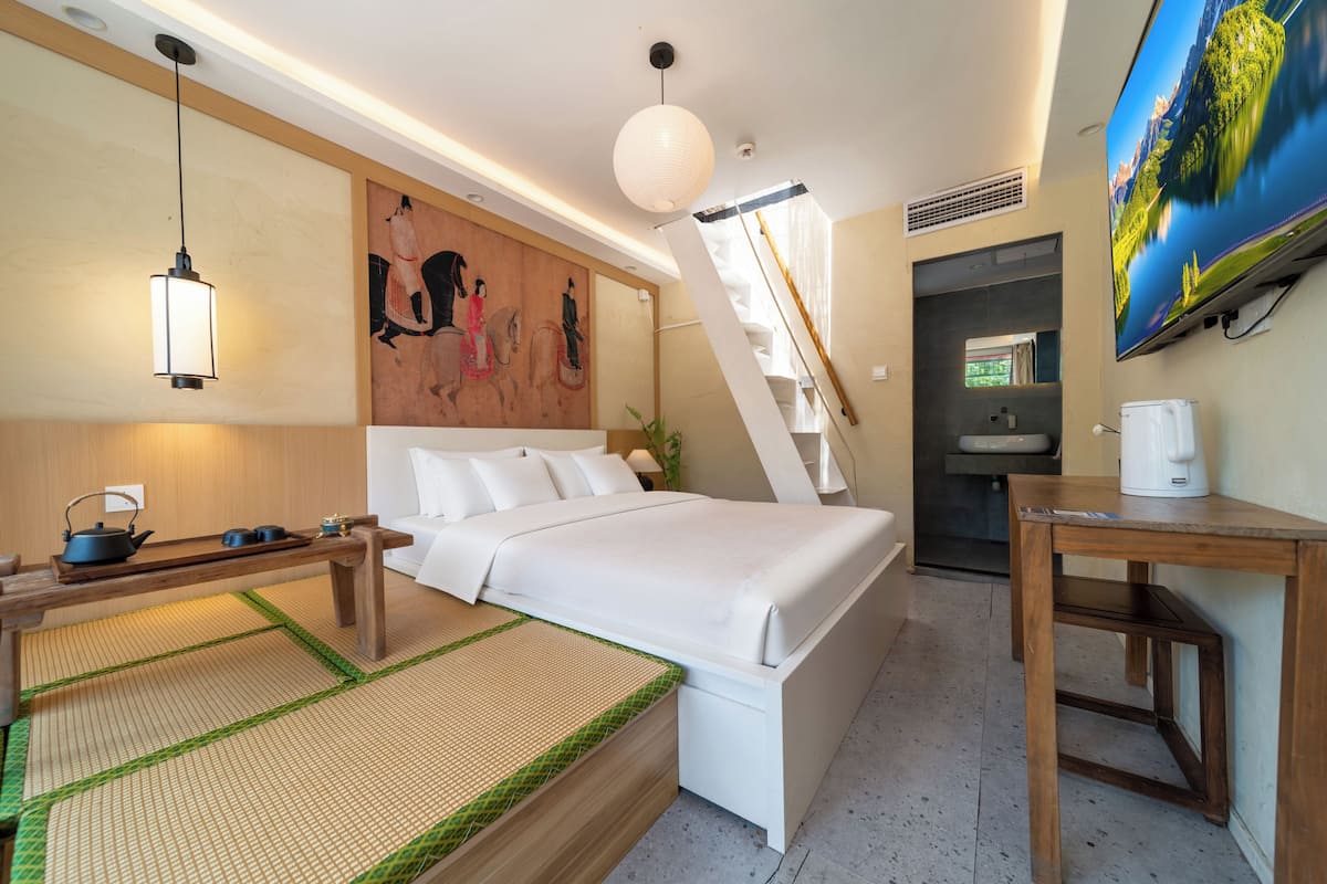A hotel room with a bed, bedside table, lamp, artwork, and a TV.