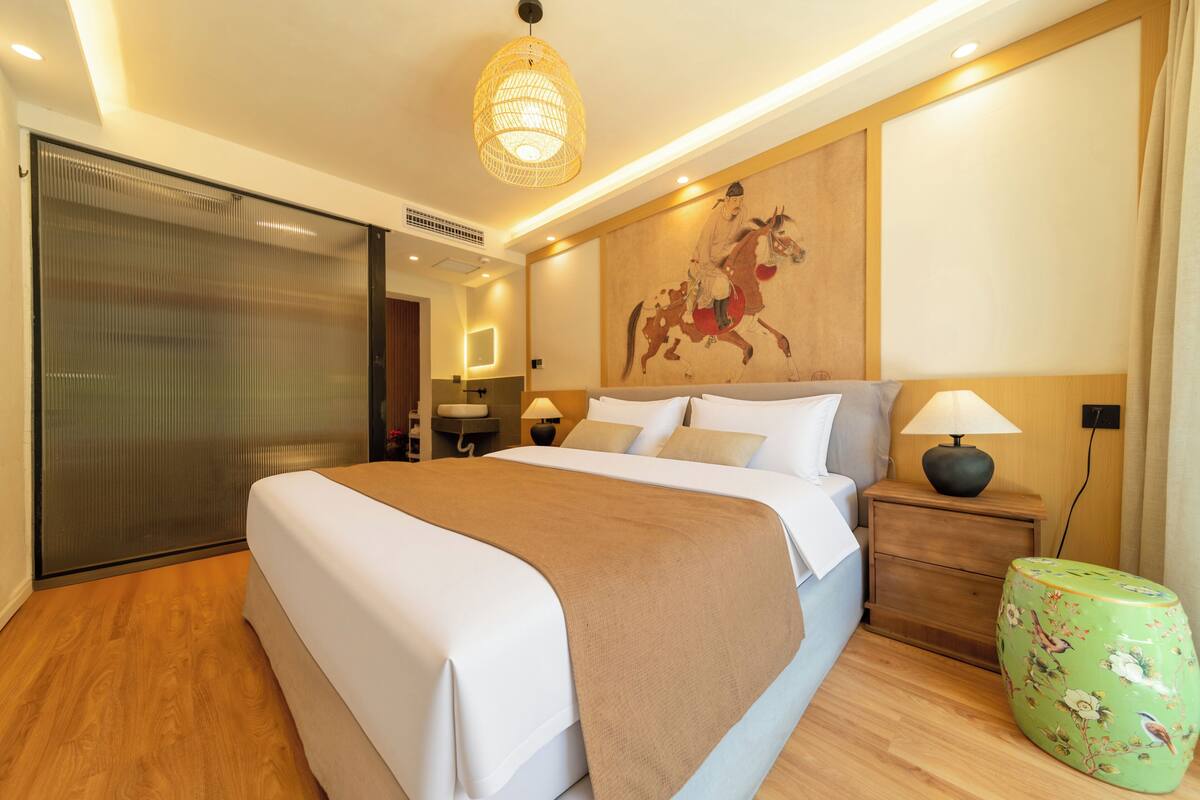 A modern hotel room with a large bed, a wooden floor, a decorative wall painting, a green stool, and a nightstand with a lamp.
