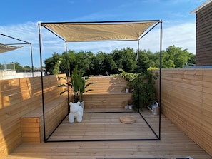 Um deck de madeira com toldo, plantas em vasos e uma pequena escultura branca.