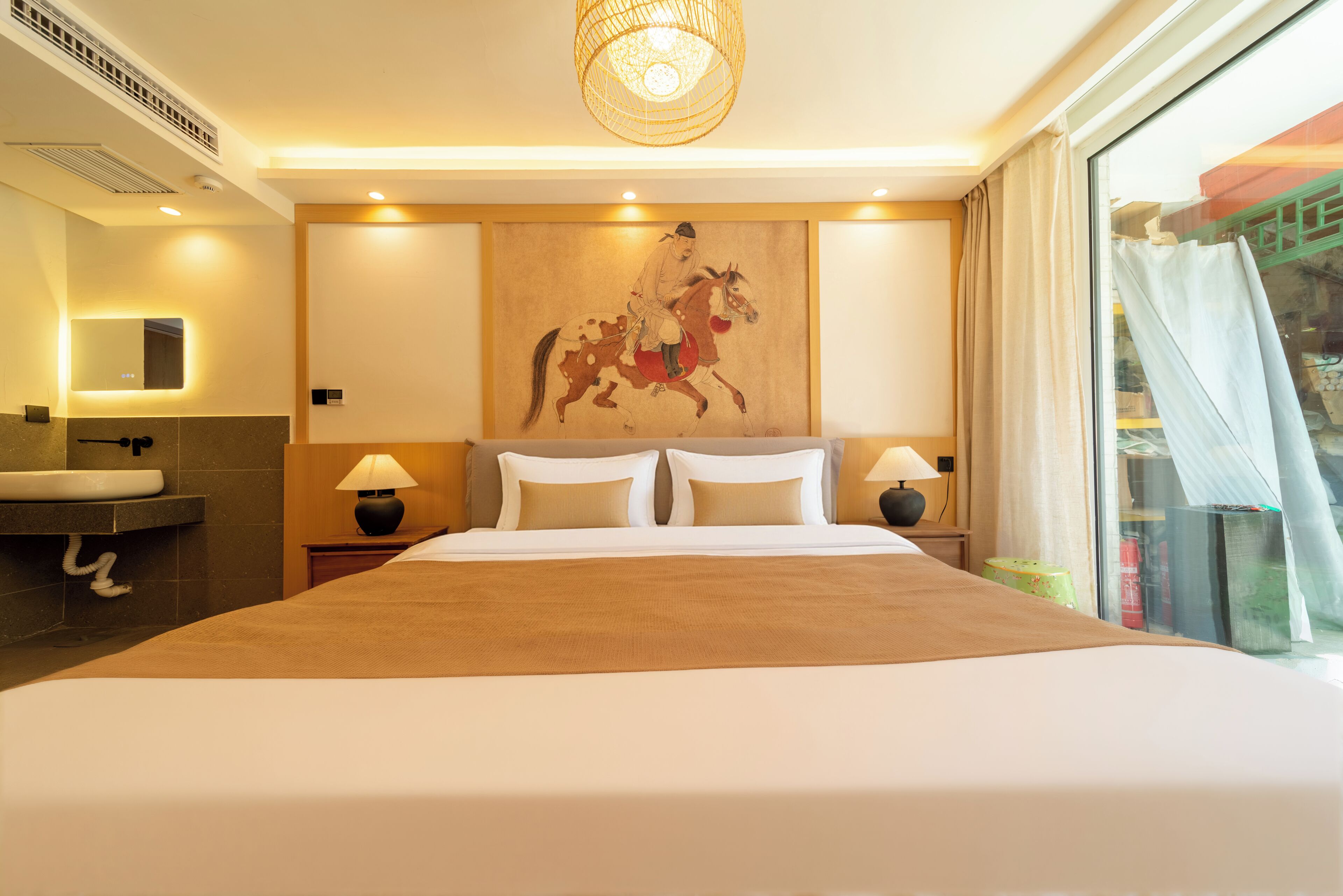 Une chambre d’hôtel avec un grand lit, deux tables de chevet, un tableau accroché au mur, une fenêtre avec des rideaux et une salle de bain équipée d’un lavabo et d’un miroir.