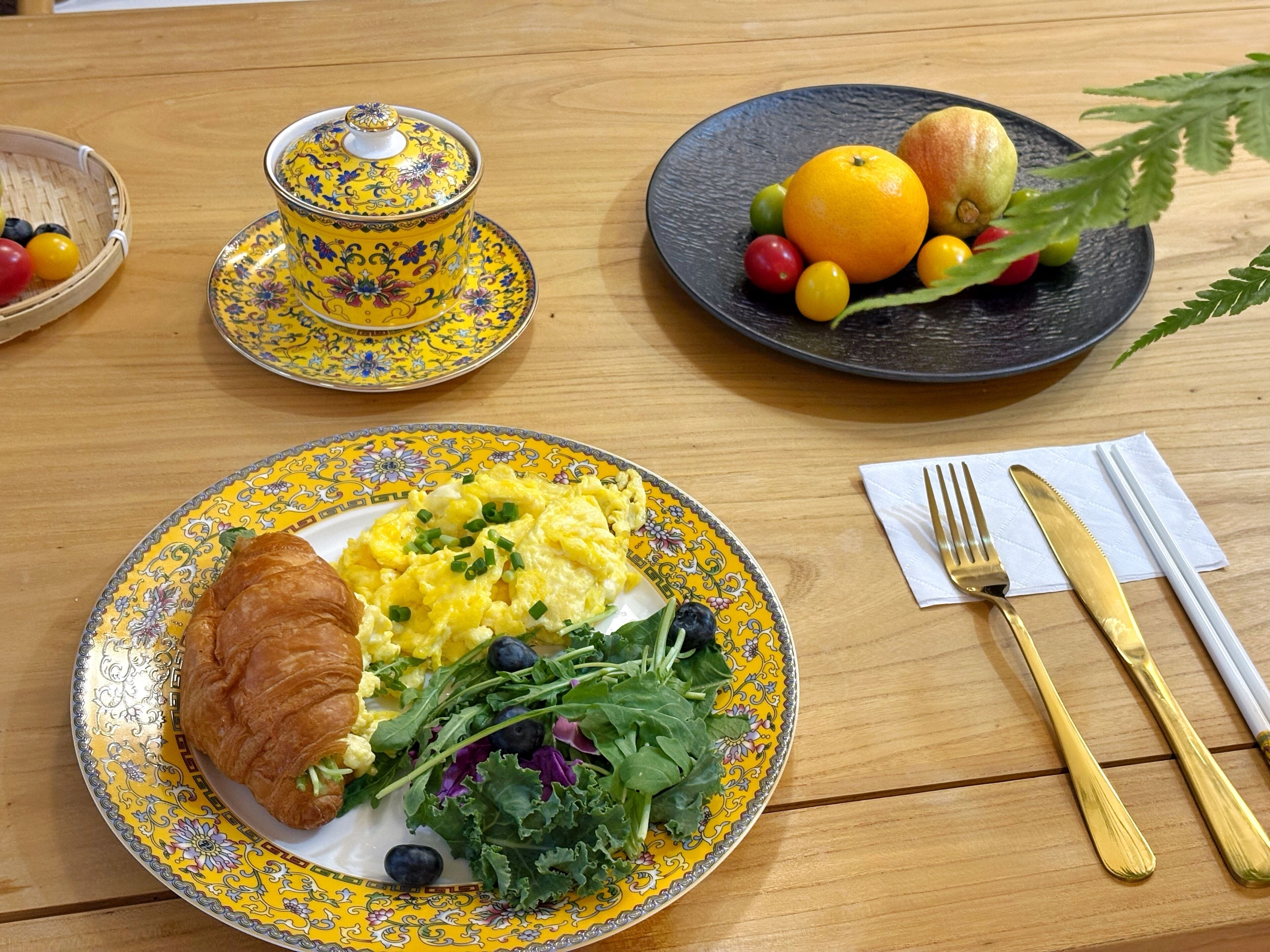 Une table en bois dressée pour le petit-déjeuner, avec une assiette d’œufs brouillés, un croissant, des légumes verts, un saladier de fruits et une théière.