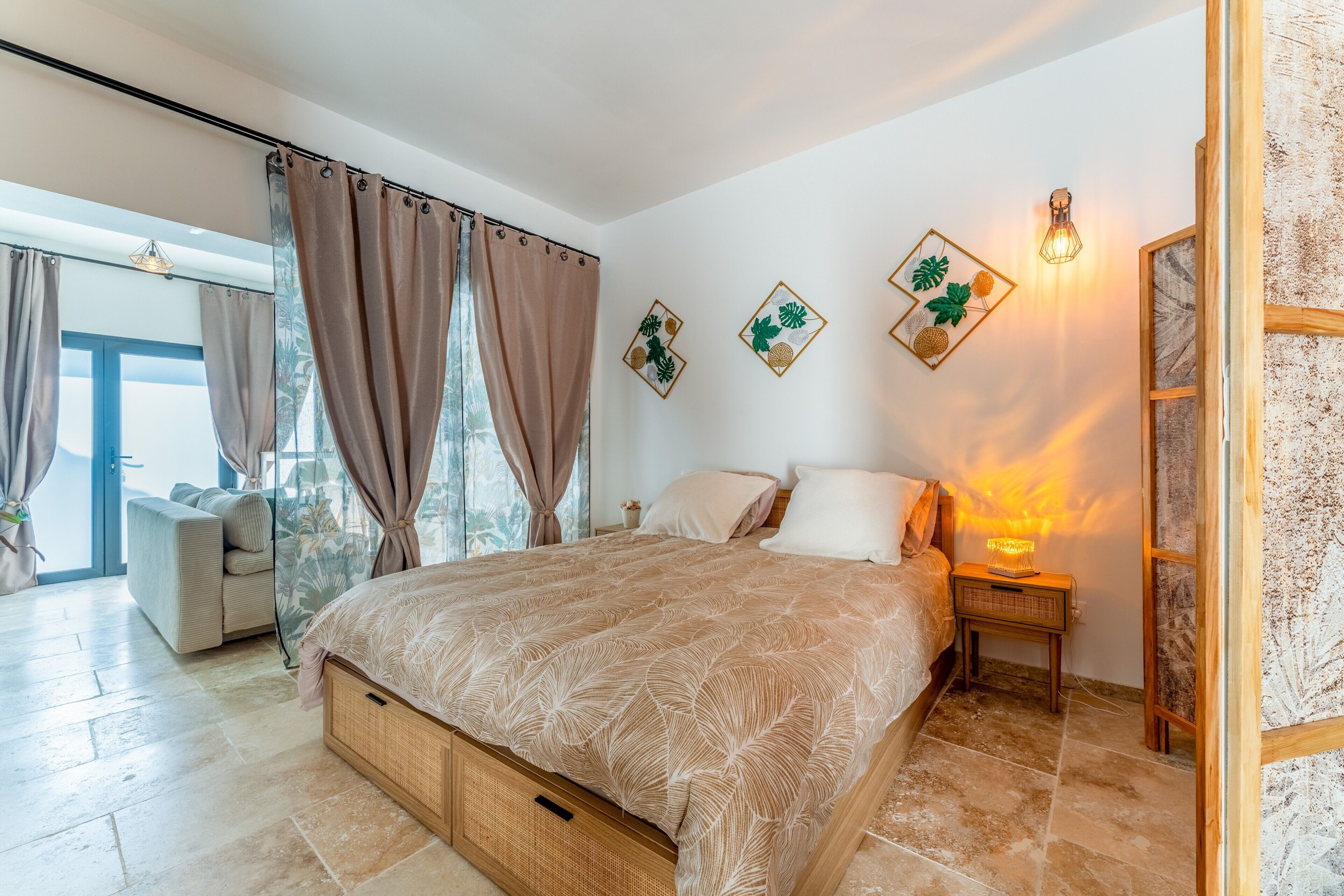 A bedroom with a bed, curtains, wall art, a nightstand, and a view of the outdoors.