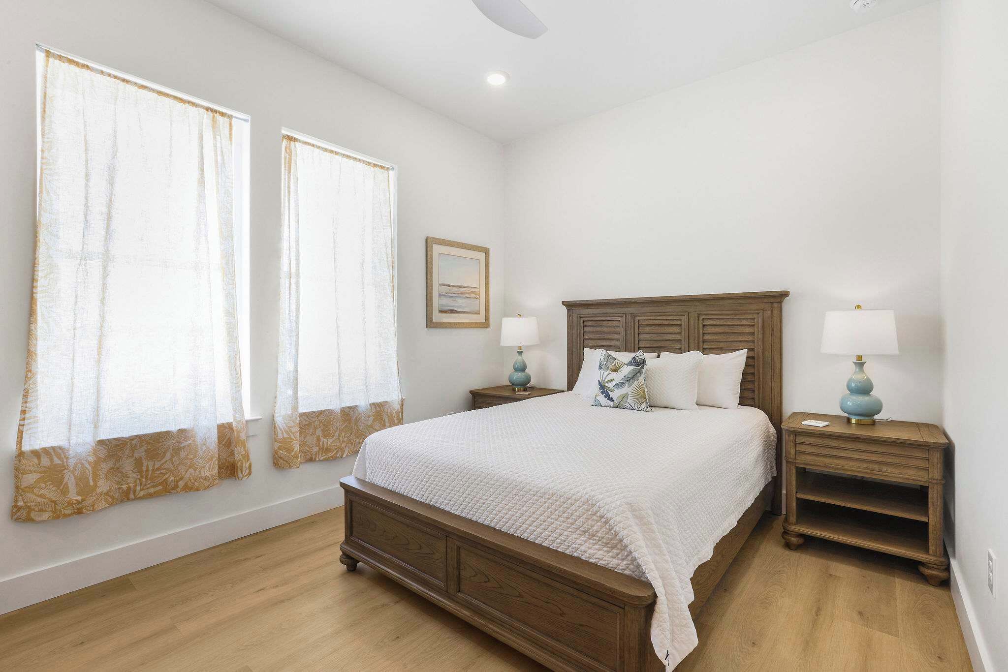 Une chambre à coucher avec un lit en bois, deux tables de chevet avec des lampes, un tableau encadré au mur et de grandes fenêtres avec des rideaux légers.