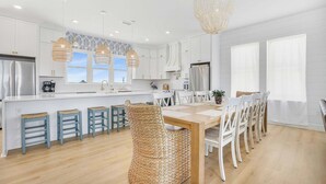 A modern kitchen with white cabinetry, a wooden dining table, and wicker bar stools. - Beach Blessing a designer coastal beach home with private pool & elevator (Gulf Shores)