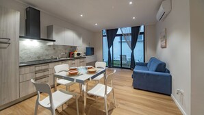 A modern kitchen with white cabinets, a dining table with chairs, and a blue sofa in a living area with large windows. - Sun & moon house (Cagliari)