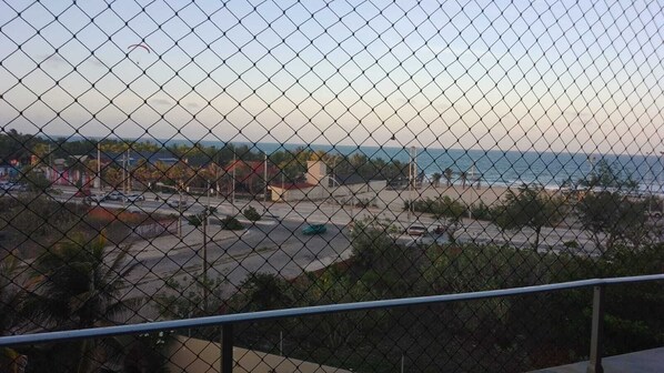 A view through a chain-link fence of a coastal area with a beach, a parking lot, and some buildings in the background.