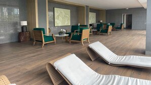 A modern lounge area with wicker lounge chairs, green upholstered chairs, and a central table with a lamp and flowers.