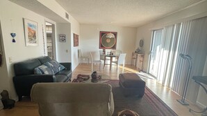 A living room with a sofa, a coffee table, and a dining table with chairs. There is a window with curtains and a door leading to another room.