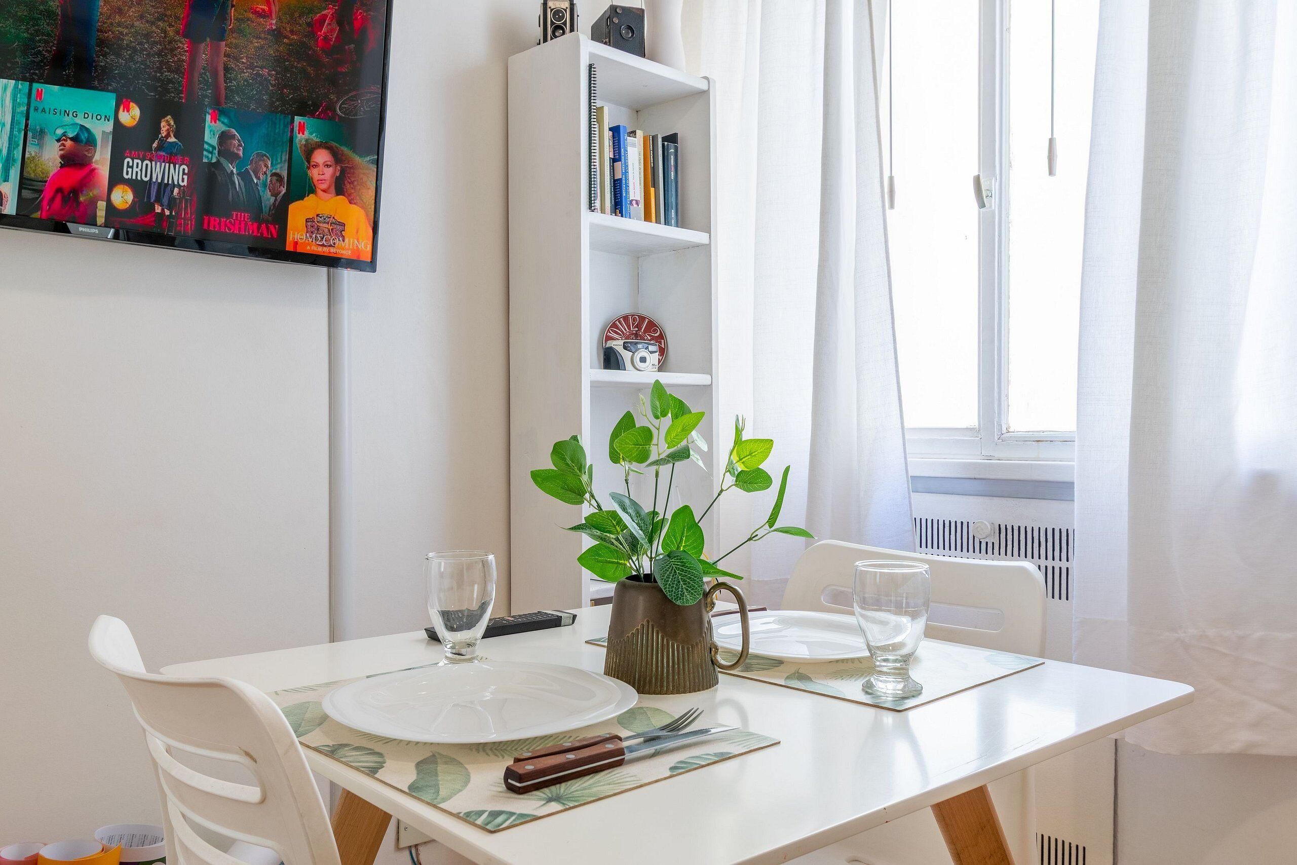 Un coin repas avec une table dressée pour deux personnes, une bibliothèque et un téléviseur affichant des affiches de films.