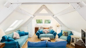 Een ruime zolderkamer met hoge plafonds, een groot raam en een gezellige zithoek met blauwe sofa’s en kussens.