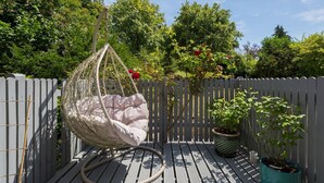 Een hangstoel met kussen op een houten terras, omringd door een witte hek en potplanten.