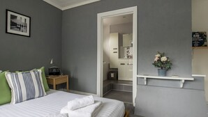 A bedroom with a bed, pillows, a bedside table with a radio, a green wall, a grey wall, a doorway to a bathroom, and a shelf with a potted plant.
