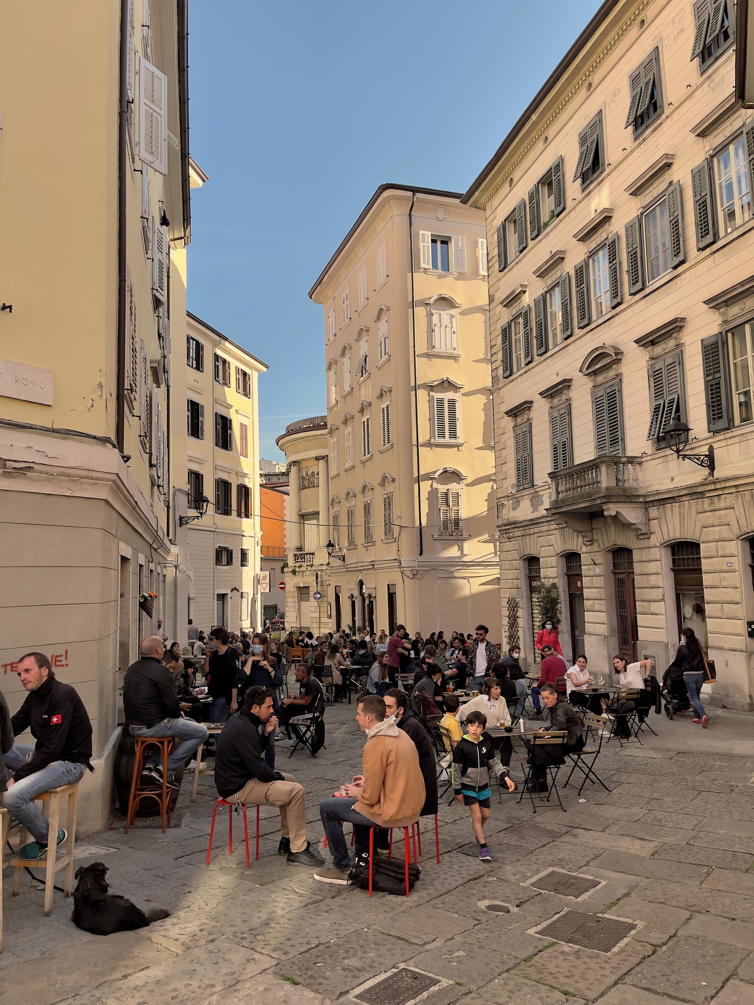 A crowded outdoor café with people sitting at tables, surrounded by historic buildings with classical architecture.