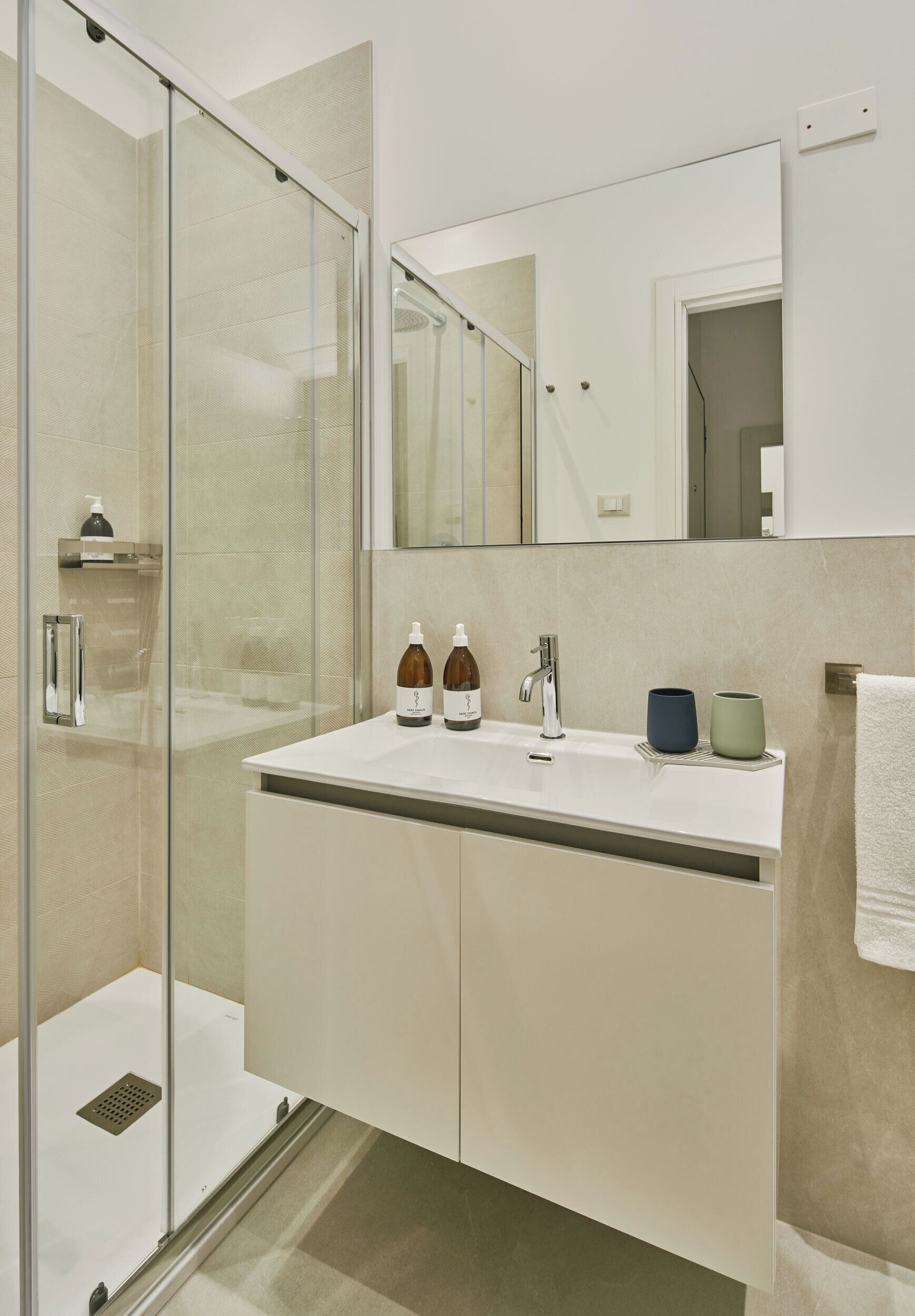 Une salle de bain moderne avec une douche à parois de verre, un lavabo blanc avec deux bouteilles et un miroir au-dessus du lavabo.