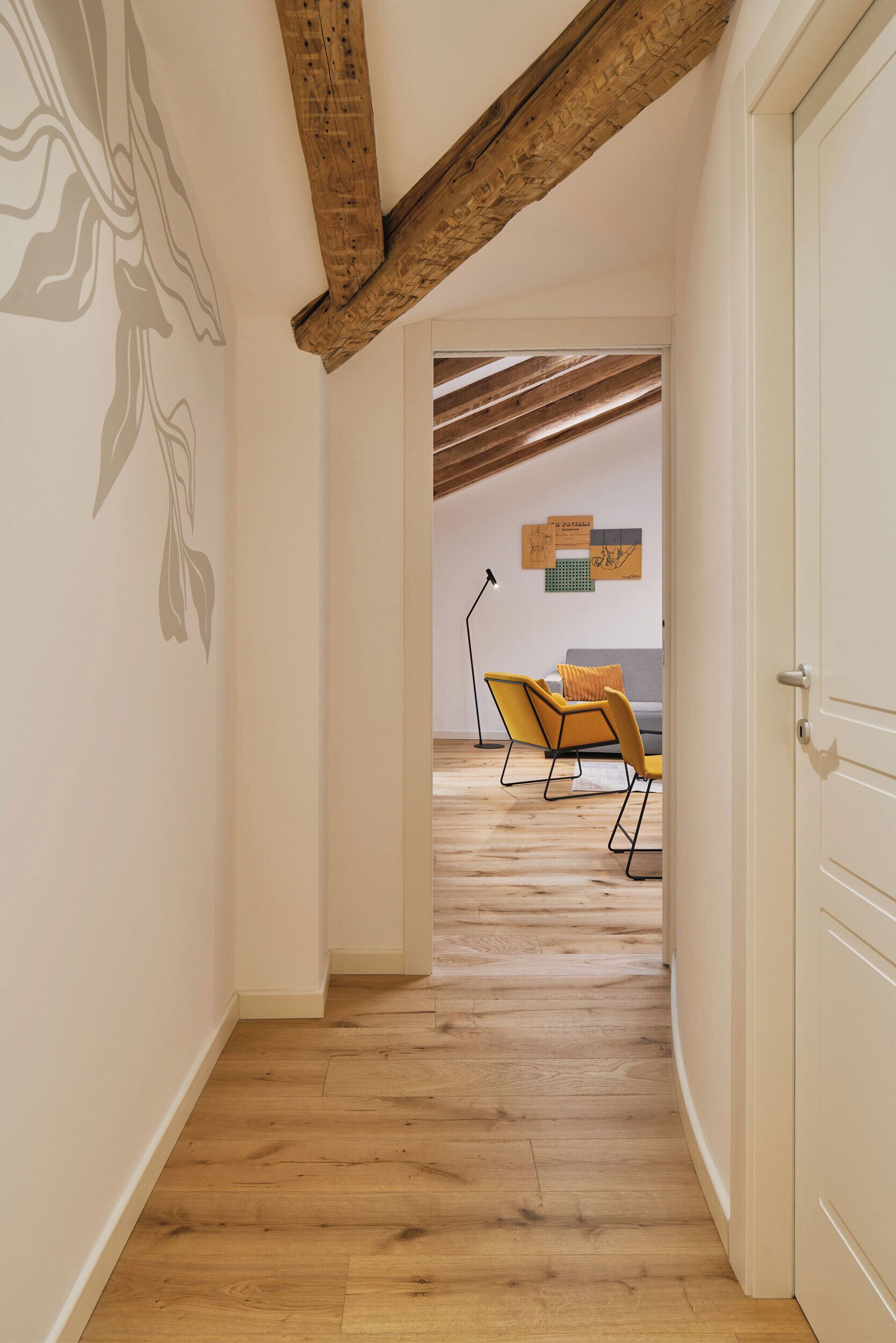 Un couloir avec un plancher en bois, une porte blanche et une pièce où l’on aperçoit un canapé et des fauteuils au fond.