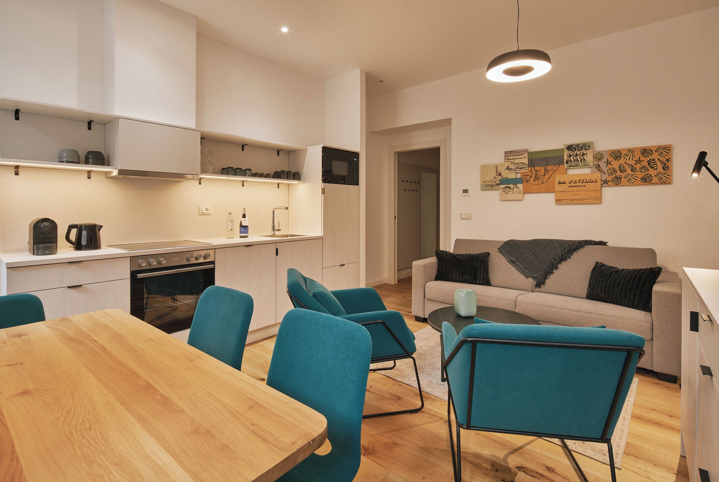 A modern kitchen with white cabinets, a wooden dining table, teal chairs, and a beige sofa in a living area.